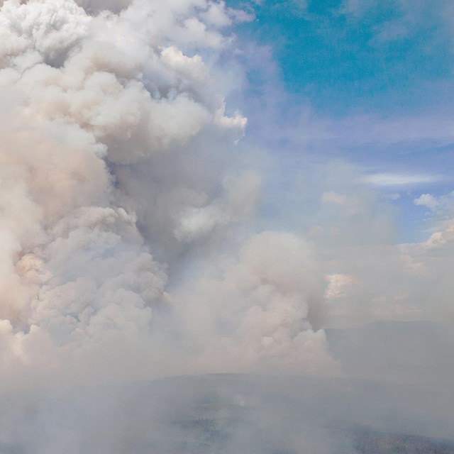 A large plume of dark, dense smoke rises into an otherwise clear, blue sky.