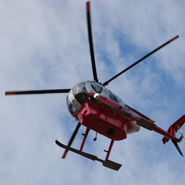 A National Park Service Huges 500 helicopter flies overhead on a sunny day.