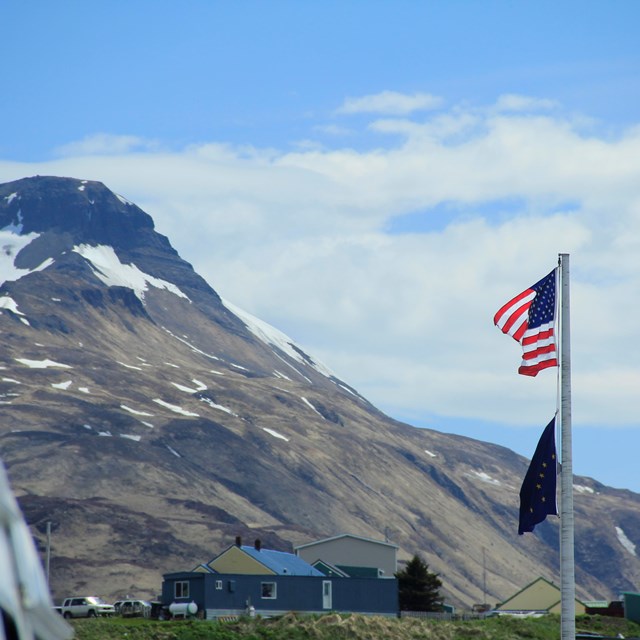 an American and Alaskan flag fly in front of a mountain peak in the Aleutian islands