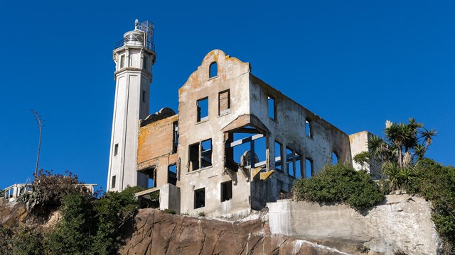 Tall lighthouse and silhouette of ruined building sit atop a cliff. 