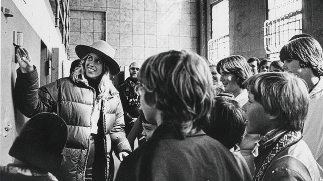 Black and white photo of a smiling female ranger with long hair and a flat hat pointing to part of t