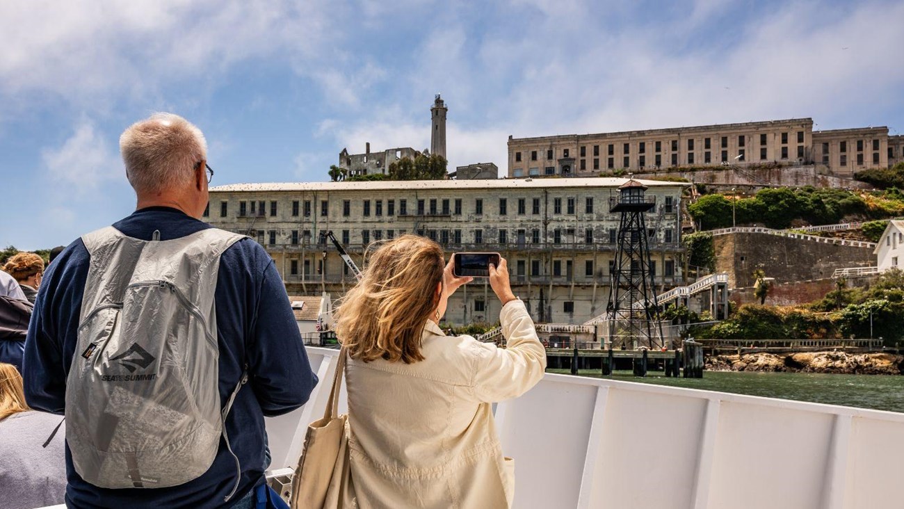 Two visitors on the bow of a boat, taking pictures of a large building on the Alcatraz dock.