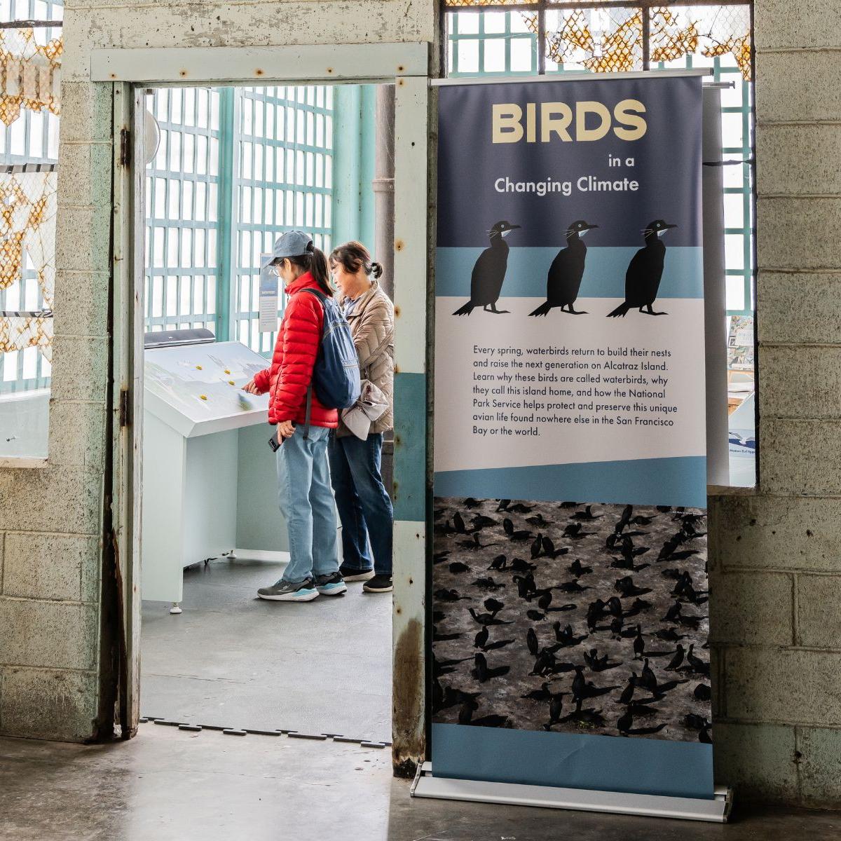 Two people stand, looking at a label in an exhibit titled 