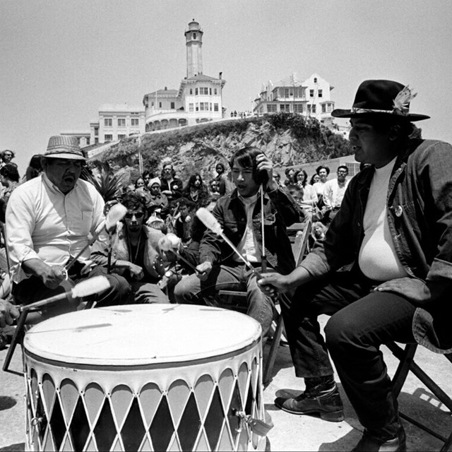 Circle of people chant and hit drum, crowd watches in background. Alcatraz buildings loom over them.