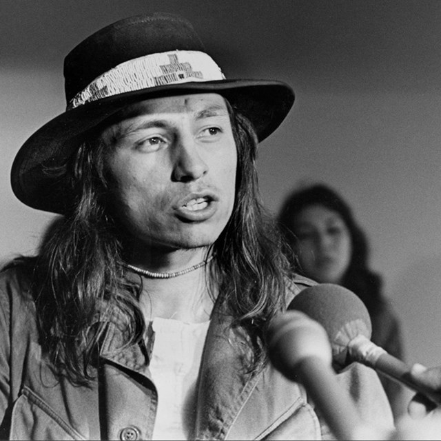 Indigenous man in wide-brimmed black hat speaks into microphone.