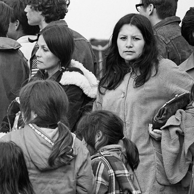 Indigenous woman stands in a crowd of people, looking directly at camera.