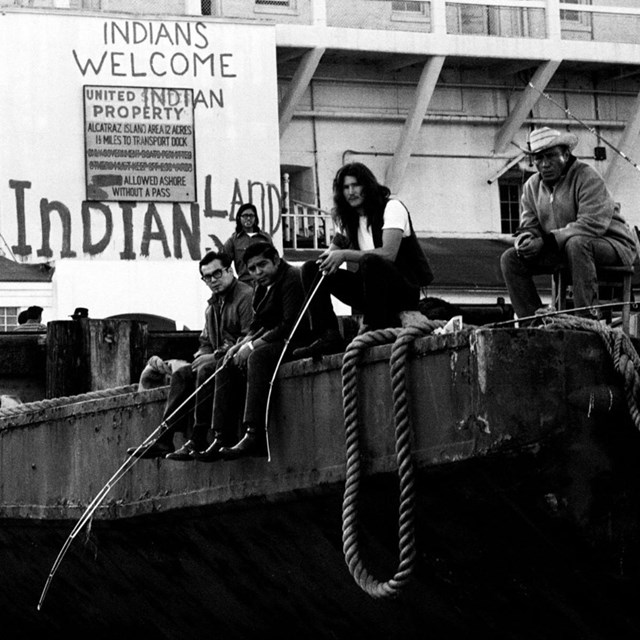 Four indigenous men sit on edge of dock, fishing. Sign behind them reads “Indians welcome,  Indian P