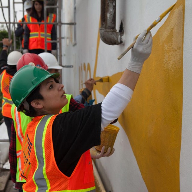 Young indigenous woman in hard hat repaints historic messaging on building with yellow paint.