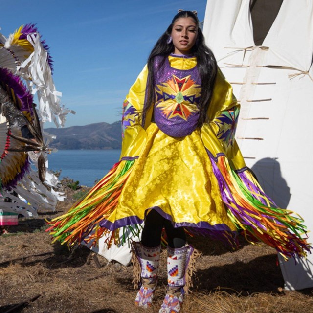 Two indigenous dancers in front of white tipi, colorful ribbons of outfits flying with movement