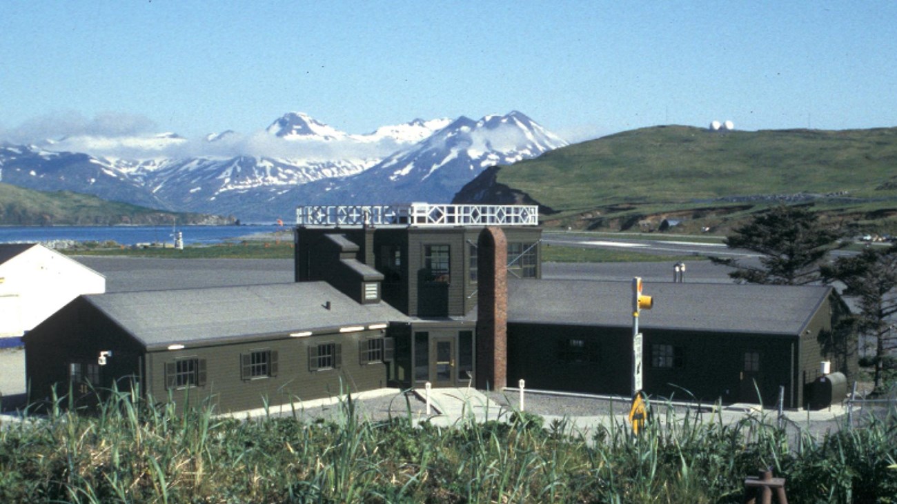 An L-shaped building with a tall lookout tower sits by a blue bay and snow-capped mountains.