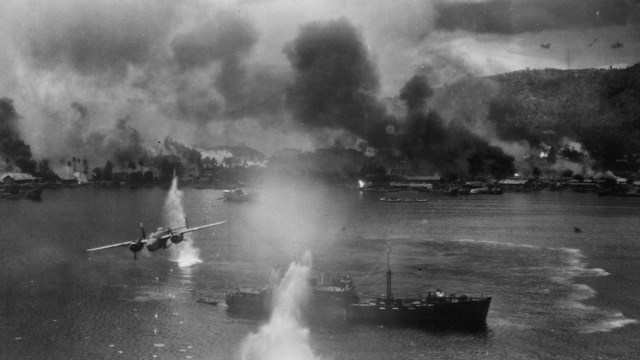 Plane flying over battleship with things exploding