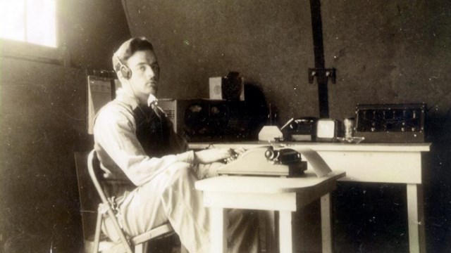 Black and white photo of man seated at typewriter, wearing radio headset, in an office. 