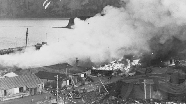 Black and white photo of a town with burning buildings and billowing smoke. 