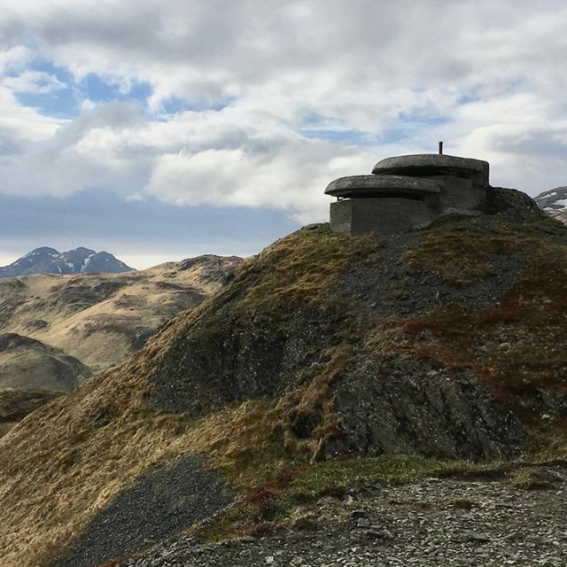 a cement historic structure sits nestled in the steep foothills of a mountain range.