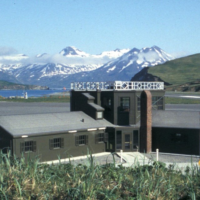 a squat, L-shaped building with a tall lookout sits by a bay surrounded by snow-capped mountaintops.