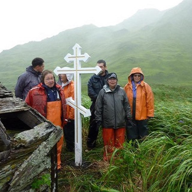 Group of people in rain great holding a white cross beside an old building in a green field. 