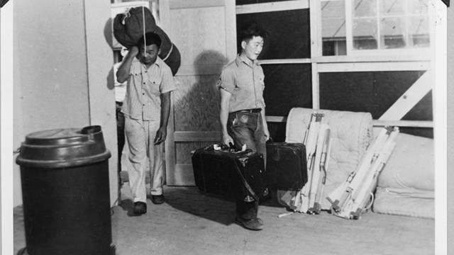 Historic image of two men walking into a barrack with two suitcases each.