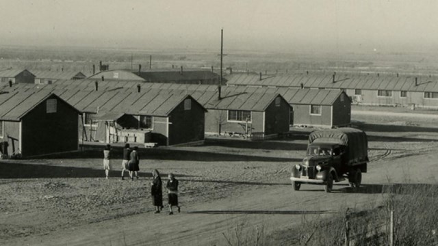 A row of army style barracks next to a road with an 1940s truck and people walking