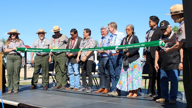 People lined up on a stage holding a ribbon for a ribbon cutting.