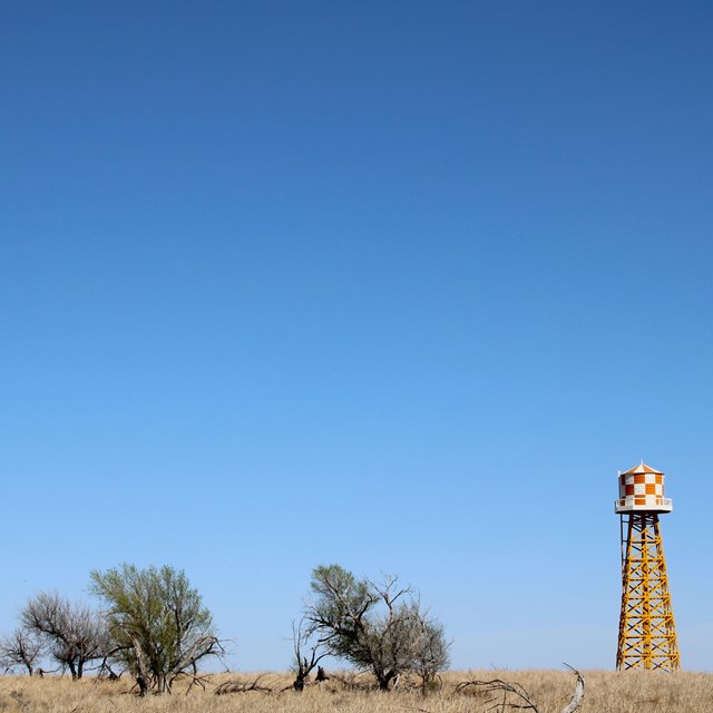 A water tower rises above a flat grassland with a few trees.