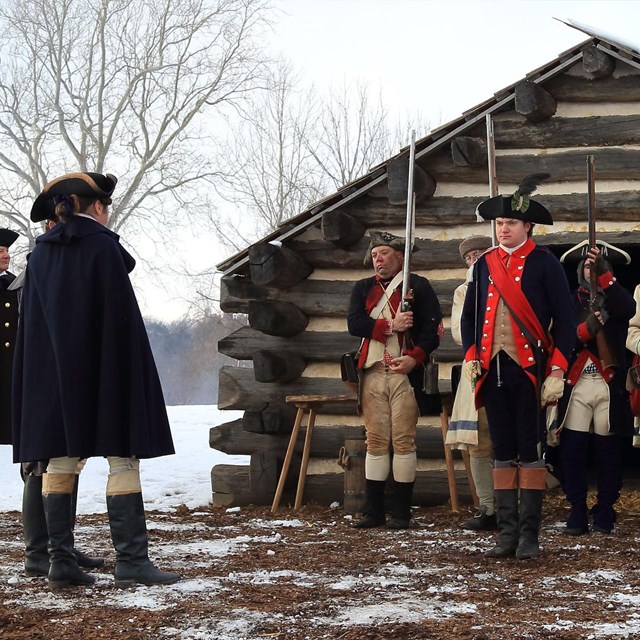Soldiers stand with their muskets presented in front of a log cabin covered with snow
