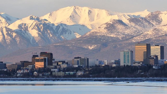 Skyline of city with snow covered mountains in background and water in foreground.