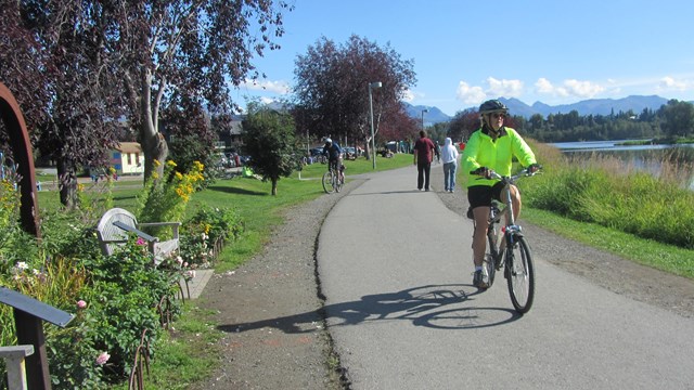 Outdoors; bike rider on a paved trail near water and greenery surrounding.