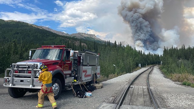 A fire truck and firefighter near train tracks with trees and smoke in the background. 