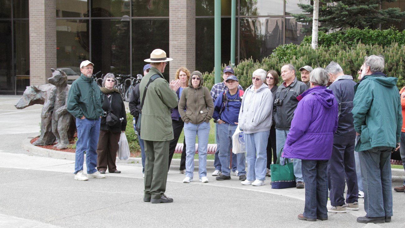 Outside on pavement with building in background, park ranger addresses a crowd of twenty people.