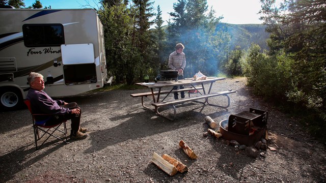 Outdoors, end of RV and trees in background, fire, picnic table, and two people in foreground.