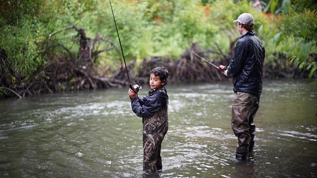Adult and child in river with hip waders and casting fishing poles.