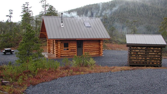 Log cabin with gravel driveway curving in front; forested outdoor setting.