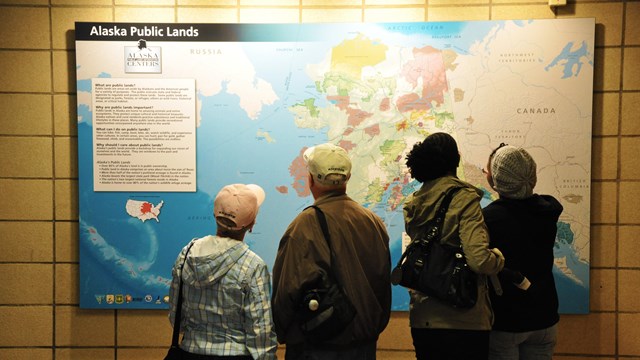 Back of four adults looking at a lit-up map on a tiled wall.