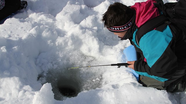 Kid laying on stomach on snow with a pole and line going in a hole.