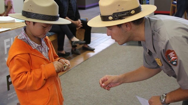 Adult Park Ranger leaning to level of child dress in ranger hat; indoors