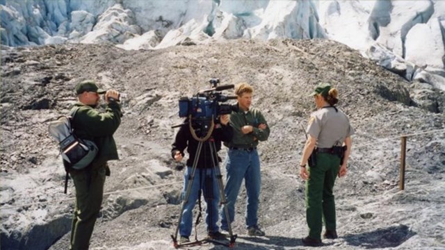 Outdoors, grey stone and ice in background; film crew with camera looing at Park Ranger.
