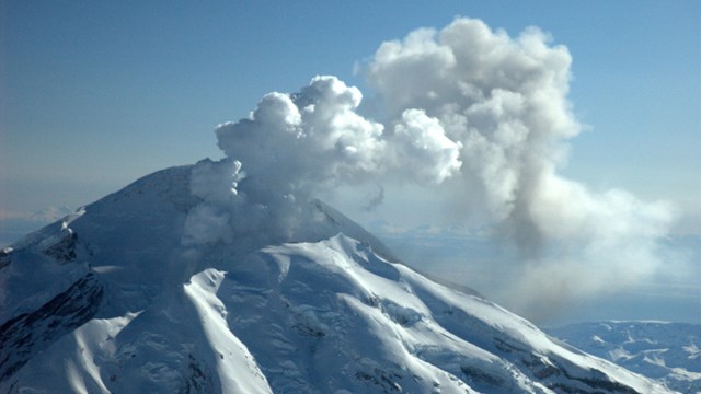 Blue sky, tall snowcapped mountain with a white cloud of steam/smoke coming out of the top.
