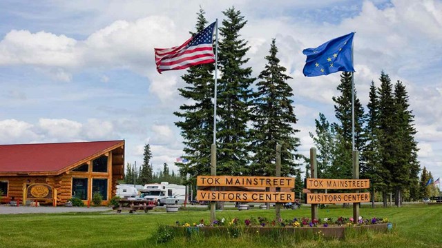 Outdoors; wooden building to left, 2 flags & 4 wooden signs in middle foreground, blue sky & grass.