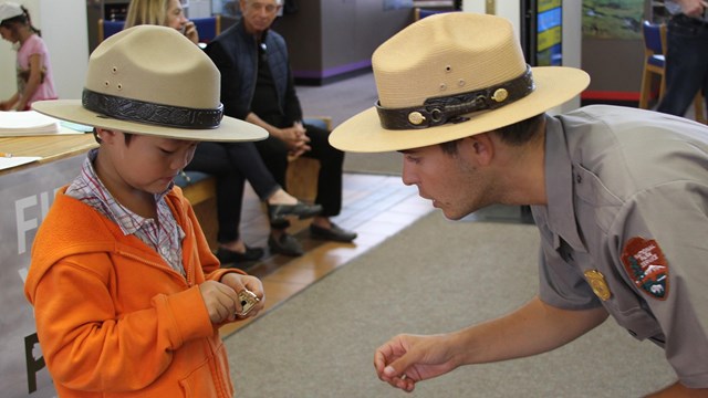 Park ranger leaning over and presenting a badge to a child in a ranger hat.