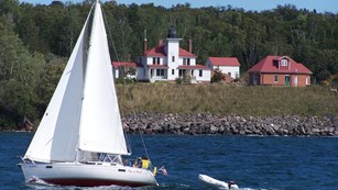 Sailboat with sails up passing in front of white lighthouse on an island cliff.