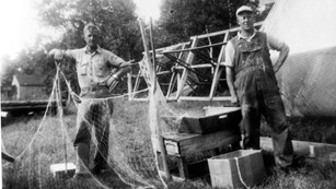A black and white photo of two men standing next their fishing nets on land. 