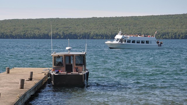 A small boat is tied to a dock, while a larger tour boat passes by.