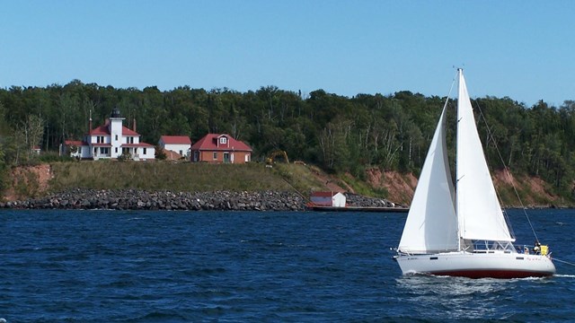 A sailboat cruises past an island with a white lighthouse up on the bluff.