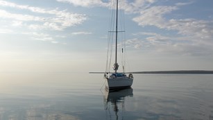 Sailboat with sails down resting on calm water with the sky and clouds reflecting off the surface. 