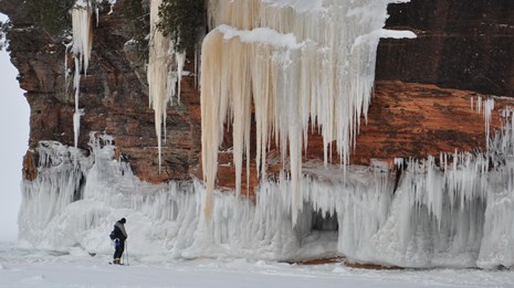 A lone person stands on a frozen lake looking at an ice-covered, sandstone cliff. 