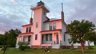 A two story white lighthouse with a red roof in a grass lawn glowing pink at sunset. 