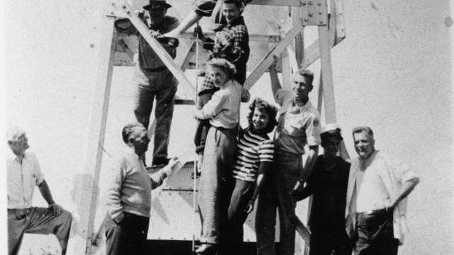 A black and white photo of a group of young adults hanging on the metal framework of a lighthouse. 