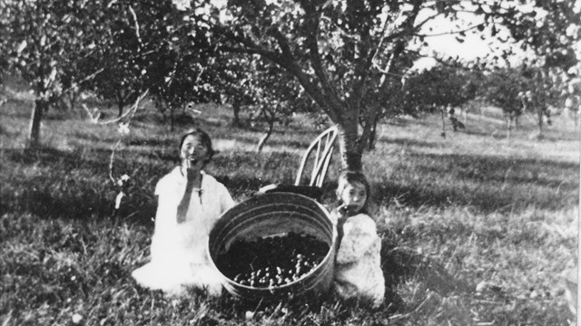 A black and white photo of two children sitting in an apple orchard and a bushel of apples. 