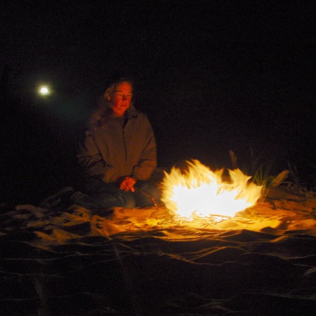 Moon shines over the shoulder of a person illuminated by a campfire. 