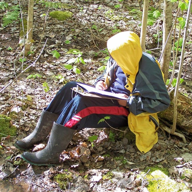 A child sits in the woods writing in a book.